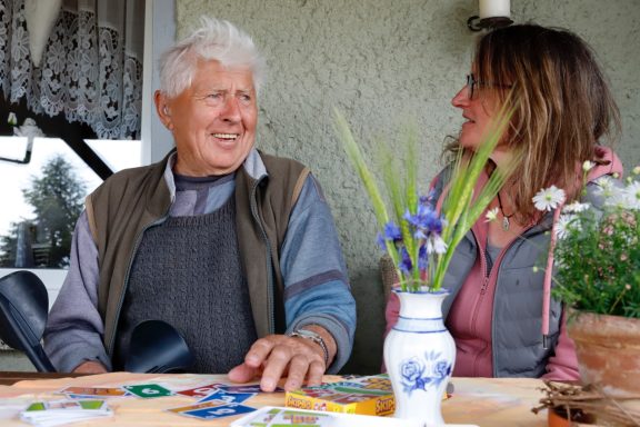 Älterer Mann und Frau im Gespräch, umgeben von Pflanzen und Karten auf dem Tisch.