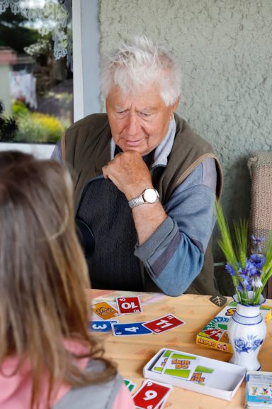 Ein älterer Mann spielt ein Kartenspiel mit einem jüngeren Mädchen. Tisch mit Karten sichtbar.