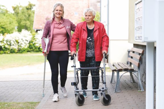 Zwei Frauen gehen gemeinsam, eine mit einem Rollator, in einer hellen Umgebung.