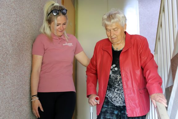 Junge Frau hilft älterer Frau beim Treppensteigen. Beide lächeln.