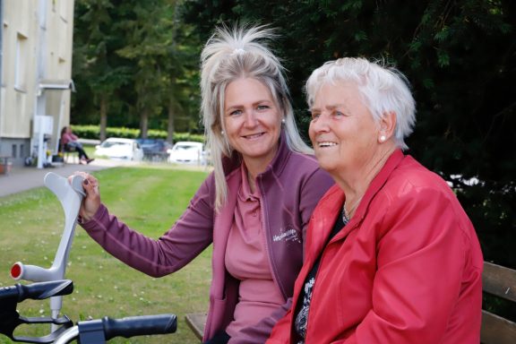 Zwei Frauen sitzen im Freien auf einer Bank, lächeln und genießen die Zeit zusammen.