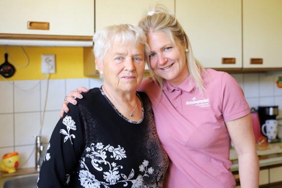 Ältere Frau und junge Frau stehen in einer Küche, beide lächeln freundlich.