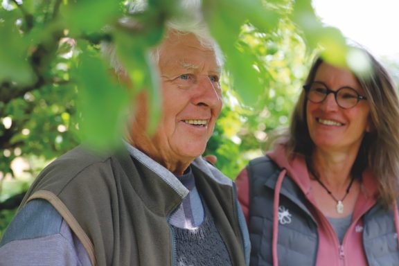 Älterer Mann und Frau stehen unter Bäumen, lächeln und genießen die Zeit zusammen.