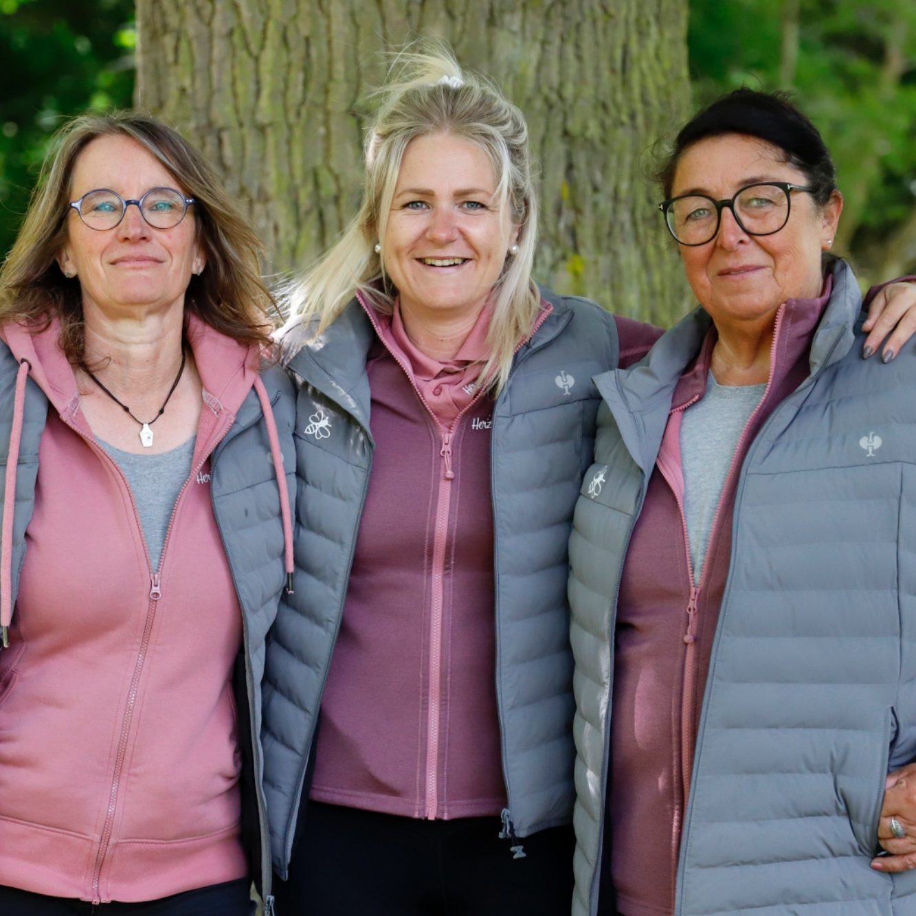 Drei Frauen stehen lächelnd vor einem Baum, tragen graue Westen und rosa Oberteile.