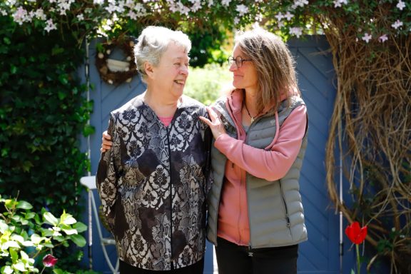 Zwei Frauen lächelnd im Garten, eine ältere Frau und eine jüngere, in enger Umarmung.